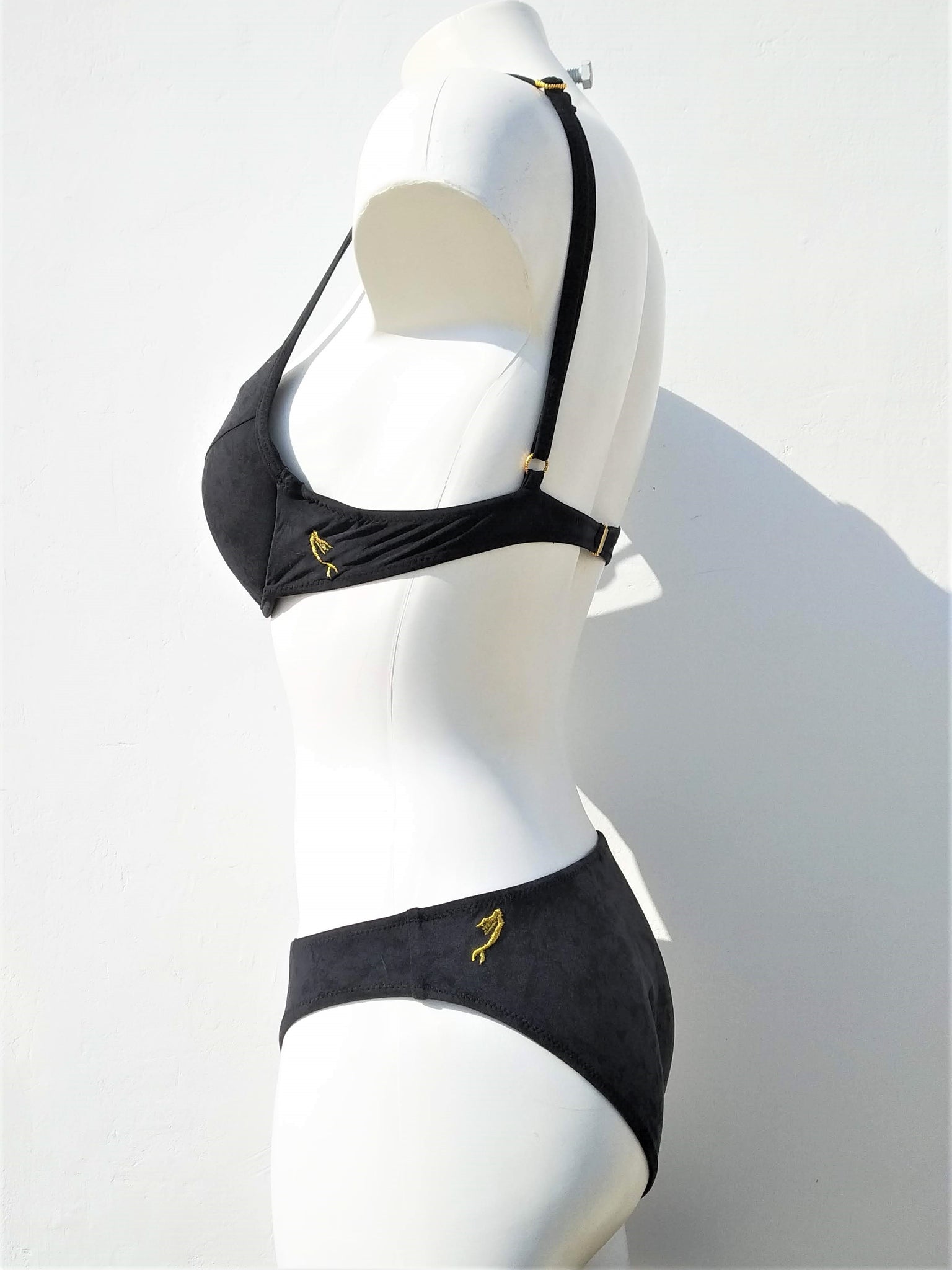 Side view of Black bikini with double push-up bra, spaghetti straps and regular black bottom, with little mermaid embroidered in gold thread, as logo, on the side of the back band of the bra, and on the side of the back of the panties.. bikinn.com